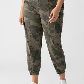 A person is wearing the Rebel Pant Hiker Camo from Sanctuary Clothing's Inclusive Collection, featuring elasticated cuffs, paired with a black top. They are also sporting white sneakers with a light-colored "V" logo on the side. The person is standing against a plain white background.