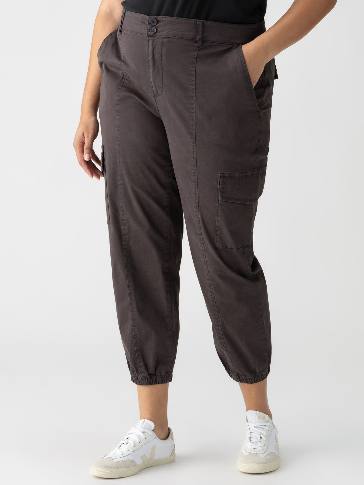 A person wearing a black top and the Rebel Pant Obsidian from Sanctuary Clothing's Inclusive Collection, featuring a high waist, side and cargo pockets, and elasticated cuffs in dark brown. The person is also wearing white sneakers with light-colored details against a plain white background.