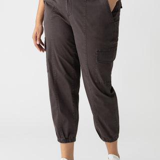 A person wearing a black top and the Rebel Pant Obsidian from Sanctuary Clothing's Inclusive Collection, featuring a high waist, side and cargo pockets, and elasticated cuffs in dark brown. The person is also wearing white sneakers with light-colored details against a plain white background.