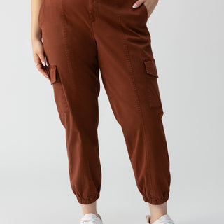 A person stands wearing Sanctuary Clothing's Rebel Pant Rich Clay from the Inclusive Collection and a patterned top. Their left hand is in a pocket, while their right arm rests down by their side. They are wearing white sneakers. The background is plain and unfocused.