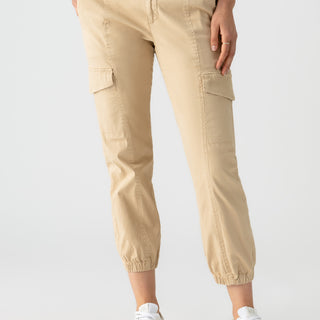 A person wearing Sanctuary Clothing's Rebel Pant in True Khaki stands against a plain white background. The pants, featuring side pockets and cuffed ankles, are paired with white sneakers. One hand is in a pocket while the person wears a partially visible white top.