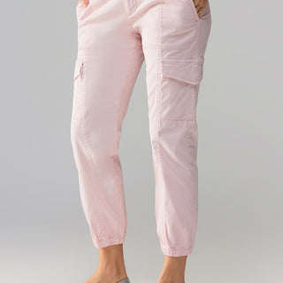 A person is dressed in Sanctuary Clothing's Rebel Pant Washed Pink No 3, featuring side pockets and gathered ankles. They are also wearing a light-colored knit sweater paired with metallic silver ballet flats. With their hands tucked into the pockets, they pose against a neutral background.