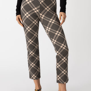 A person wearing the Carnaby Kick Crop Legging Graphic Plaid pants by Sanctuary Clothing in a black and beige plaid pattern, paired with a black long-sleeve top. The person's hands are by their sides, and they are wearing black flats with ankle straps. The background is plain white.