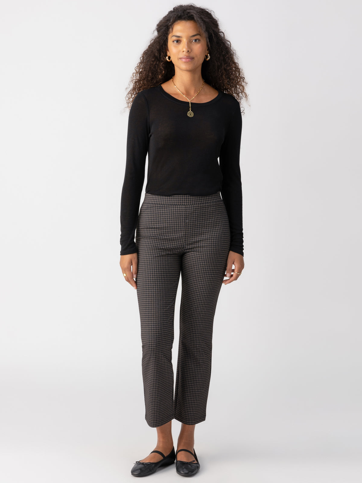 A woman with long curly hair stands against a plain white background. She wears a long-sleeve black top, Sanctuary Clothing's Carnaby Kick Crop Shadow Check pants, black flats, and a gold necklace with a circular pendant. Her hands rest by her sides, and she has a calm expression.