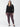 In front of a simple white backdrop, an individual is dressed in a black long-sleeve button-up shirt and Sanctuary Clothing's Runway Legging Mars Red Check from the Inclusive Collection. They complete the look with black flats.