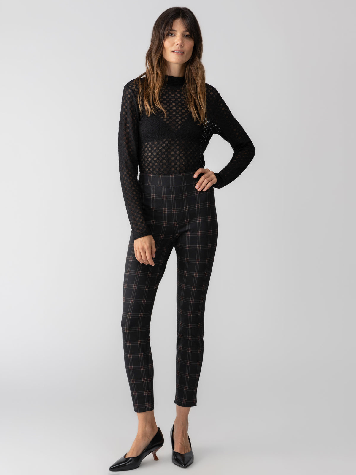 A person stands against a plain background wearing a black sheer long-sleeve top and Sanctuary Clothing's Runway Legging Garnet Plaid pants. They have long, wavy hair and are wearing black heels. They have one hand by their side and the other gently touching their hip.