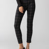 A person is wearing the Runway Legging Garnet Plaid pants from Sanctuary Clothing, paired with a black long-sleeve top featuring a mesh pattern on the sleeves. They are standing with one hand slightly touching the other, and wearing black pointed-toe heels. The background is plain light gray.