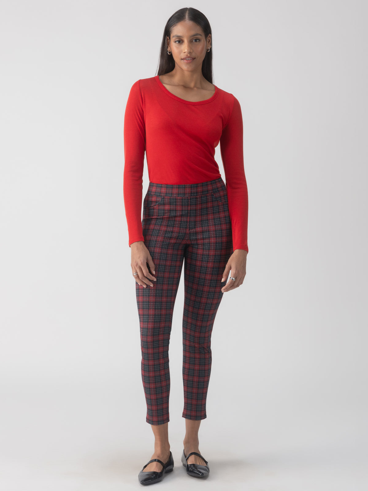 A person with long hair is wearing a red long-sleeve top and the Runway Legging Mars Red Check from Sanctuary Clothing, featuring a black, red, and gray plaid pattern. They are standing against a plain gray background and have black flat shoes on.