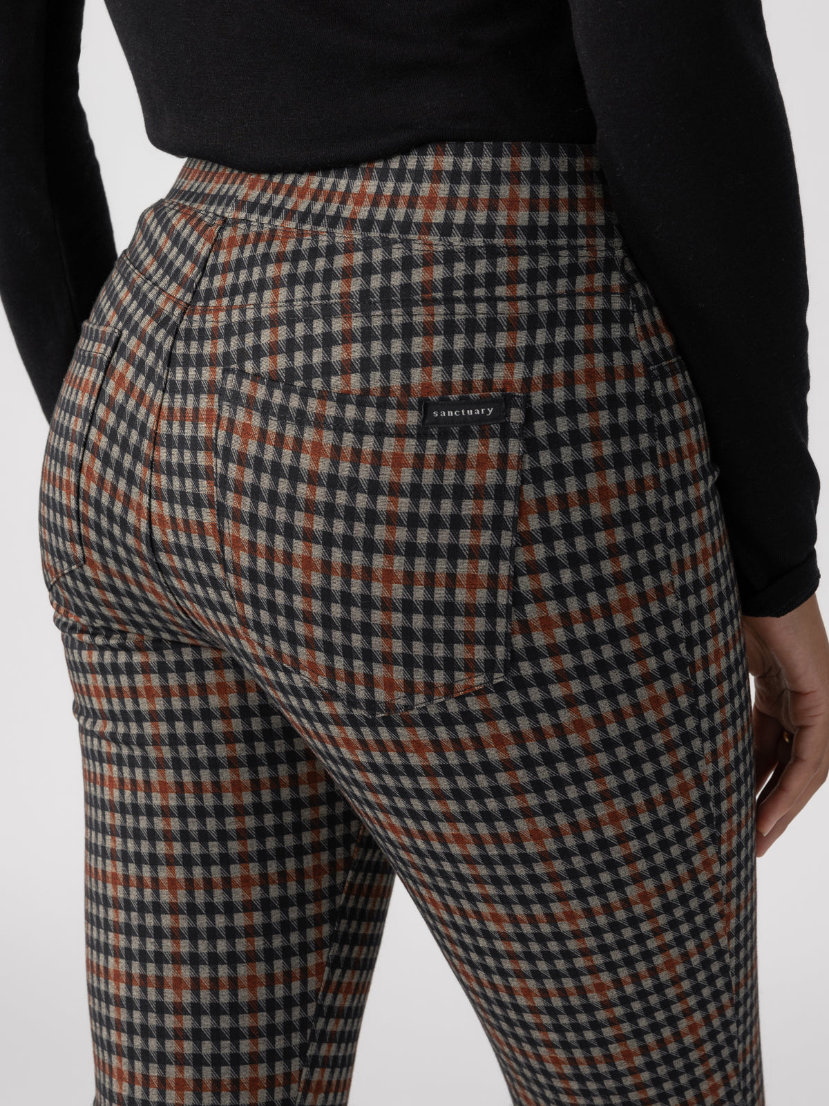 A person wearing the Runway Legging Arrow Plaid from Sanctuary Clothing and a black top is shown from the back, focusing on the hip area. The pants have visible back pockets, and the brand name, "Sanctuary," is subtly printed above the right pocket.