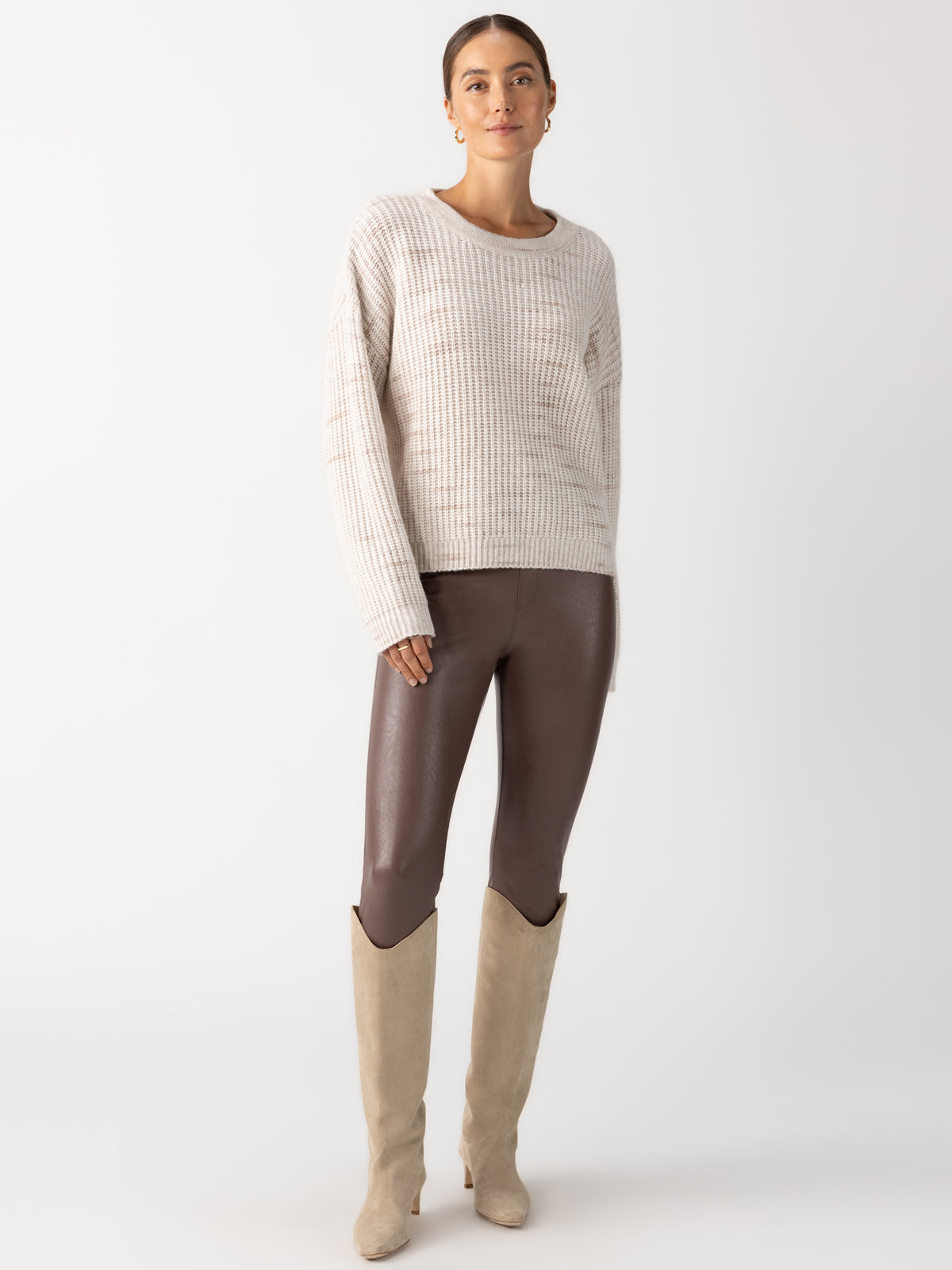 A person stands against a neutral background wearing a cream ribbed sweater, Sanctuary Clothing's Runway Legging in Dark Feather, and beige knee-high boots.