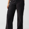 A person wearing Sanctuary Clothing's Reissue Cargo Pant Black from the Inclusive Collection, featuring a high-waisted fit with multiple pockets and a relaxed, wide-leg silhouette. They have paired it with a black-and-white printed top and are standing confidently with one hand on their hip and the other in their pocket. Completing the look, they are wearing black flats.