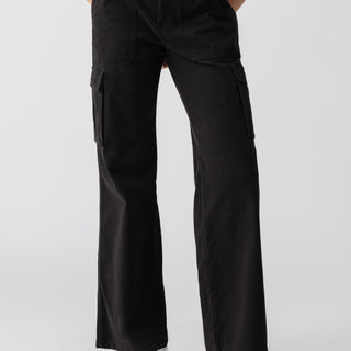 A person is wearing Sanctuary Clothing's Reissue Cargo Black wide-leg pants with large side pockets, paired with a white T-shirt. They are standing against a plain background, with hands in the pants' pockets. White sneakers with light-colored soles are visible on their feet.