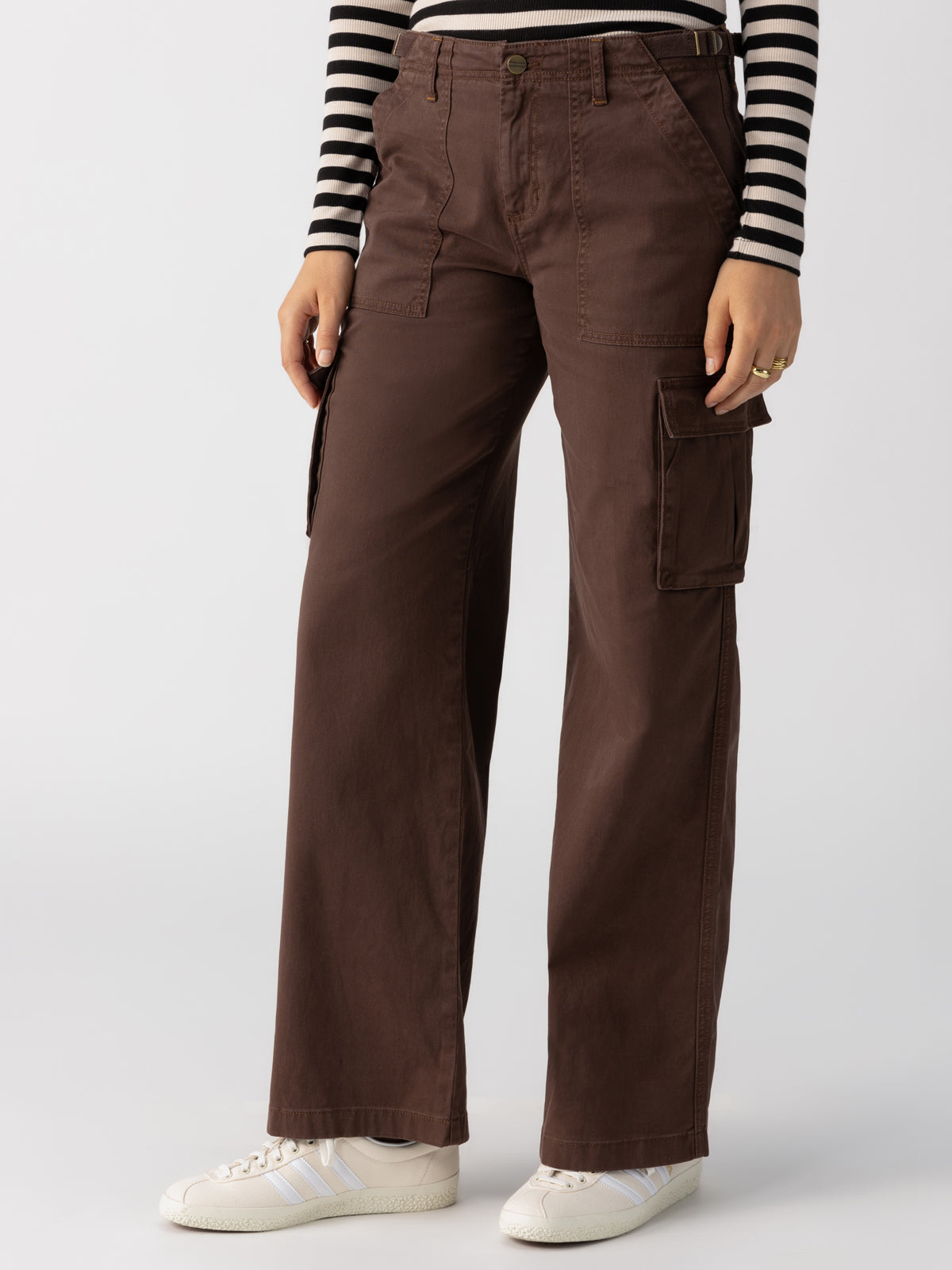 A person stands wearing the high-waisted Sanctuary Clothing Reissue Cargo Pants in Brown Sugar with multiple pockets, paired with a black and white striped long-sleeve shirt and white sneakers. The image focuses on the lower body and partial torso against a plain background.