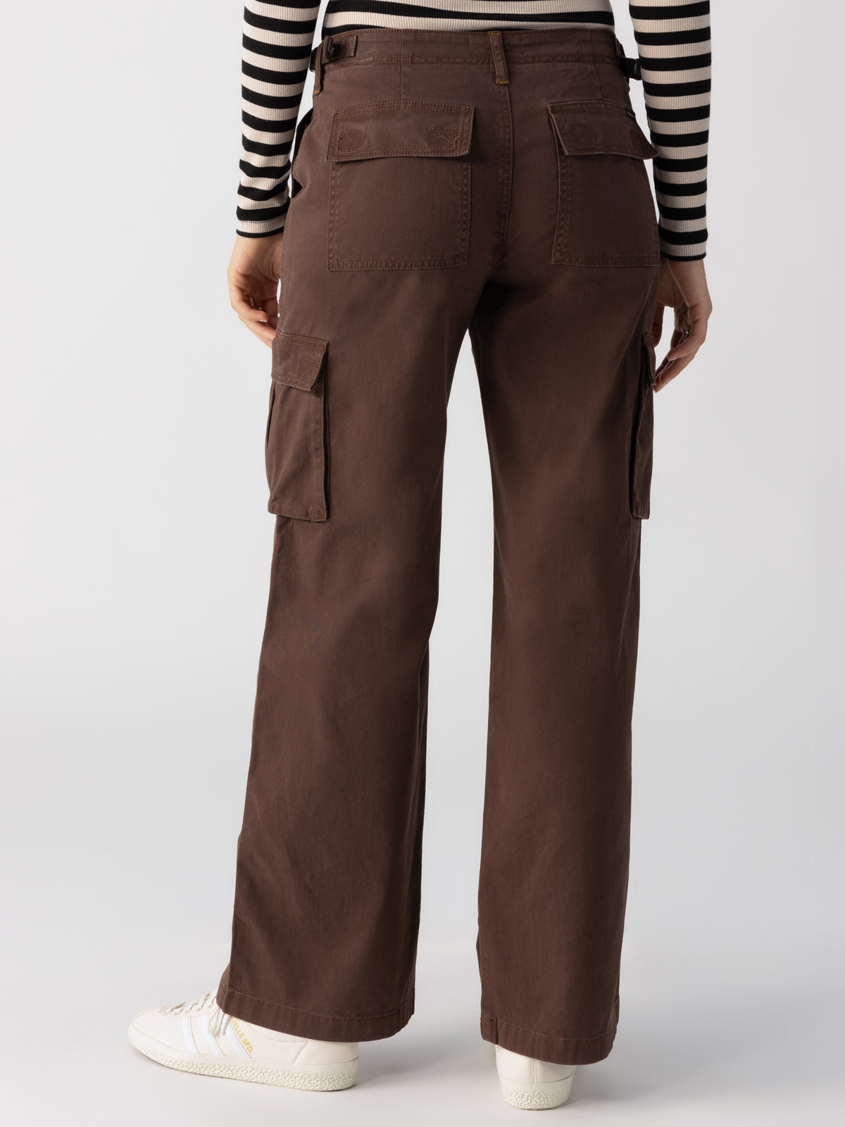 A person is shown from the back, wearing the Reissue Cargo Brown Sugar pants by Sanctuary Clothing, which feature large pockets. They also wear a black and white striped long-sleeve shirt and white sneakers. The background is a plain light-colored wall.