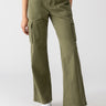 A person is wearing the Reissue Cargo Pant in Mossy Green from Sanctuary Clothing, which are high-waisted with multiple pockets, paired with a white T-shirt. They are also sporting white sneakers. The background is plain and light-colored.