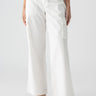 A person is dressed in Sanctuary Clothing's Reissue Cargo White pants, featuring high-waisted, wide-leg design and large thigh pockets with hands partially tucked in. They are matched with black strappy sandals and a black top peeking out from the bottom. The background is plain white.