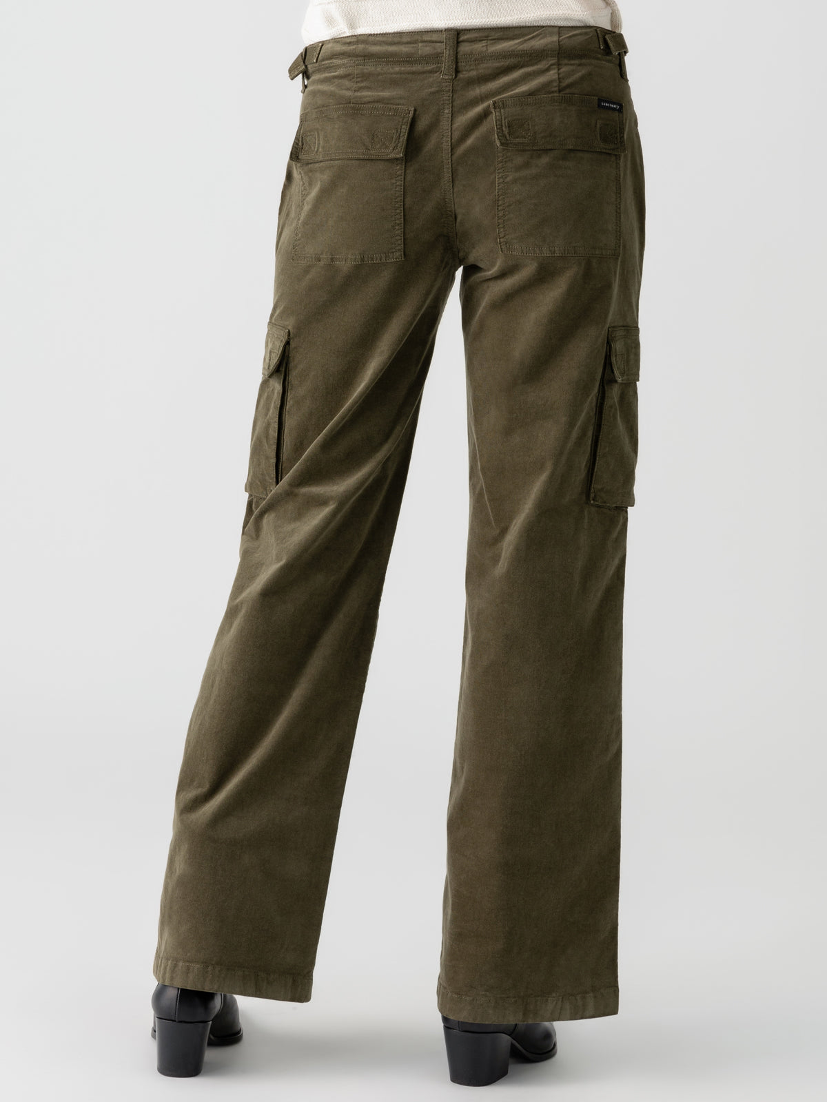 A person wearing Sanctuary Clothing's Reissue Cargo Burnt Olive pants with side pockets and black heeled shoes stands facing away from the camera. The cargo pants feature a loose, straight fit, and the person pairs them with a light-colored shirt tucked in. The background is plain and light-colored.