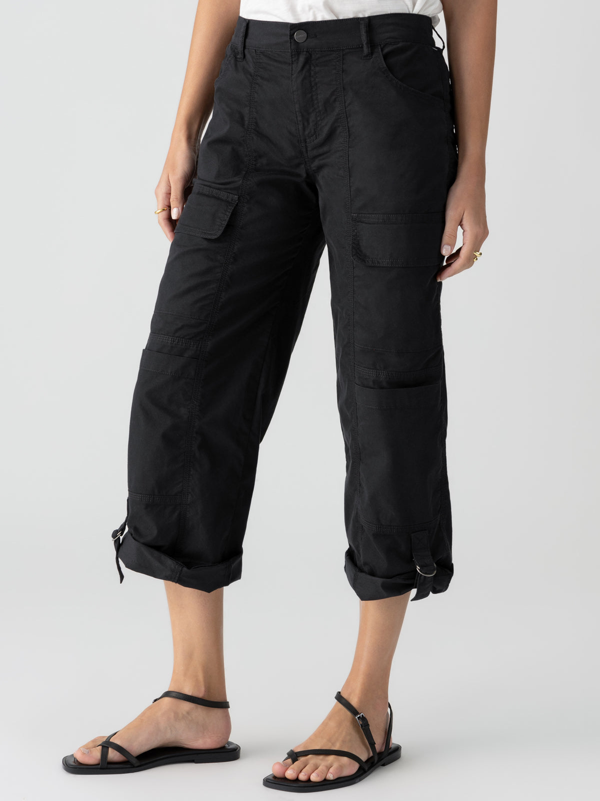 A person is wearing the Cali Cargo Pant Black by Sanctuary Clothing, featuring multiple pockets and rolled up to mid-calf length. They are also sporting black ankle-strap sandals, with a partially visible white top. The background is plain and light-colored.