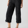 A person is wearing the Cali Cargo Pant Black by Sanctuary Clothing, featuring multiple pockets and rolled up to mid-calf length. They are also sporting black ankle-strap sandals, with a partially visible white top. The background is plain and light-colored.