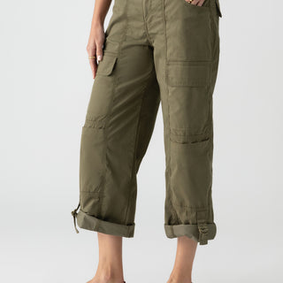 Person wearing Sanctuary Clothing's Cali Cargo Pant in Mossy Green, which features multiple pockets and rolled-up cuffs, paired with tan flat sandals. The person’s hand is resting in their pocket, and they are standing against a plain background. Only the lower half of the body is visible.