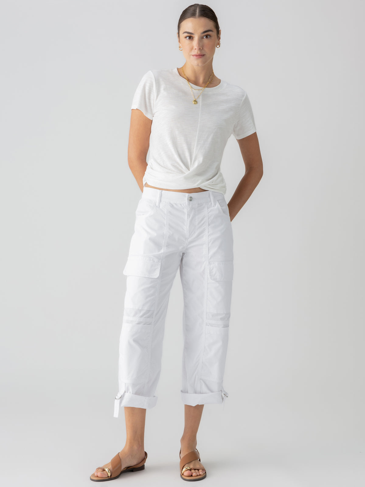 A person stands against a plain background, wearing a white short-sleeve t-shirt and the Cali Cargo Pant in White by Sanctuary Clothing, with the pants stylishly rolled up at the ankles. They also sport brown sandals and a necklace, with their hands casually placed in their pockets.