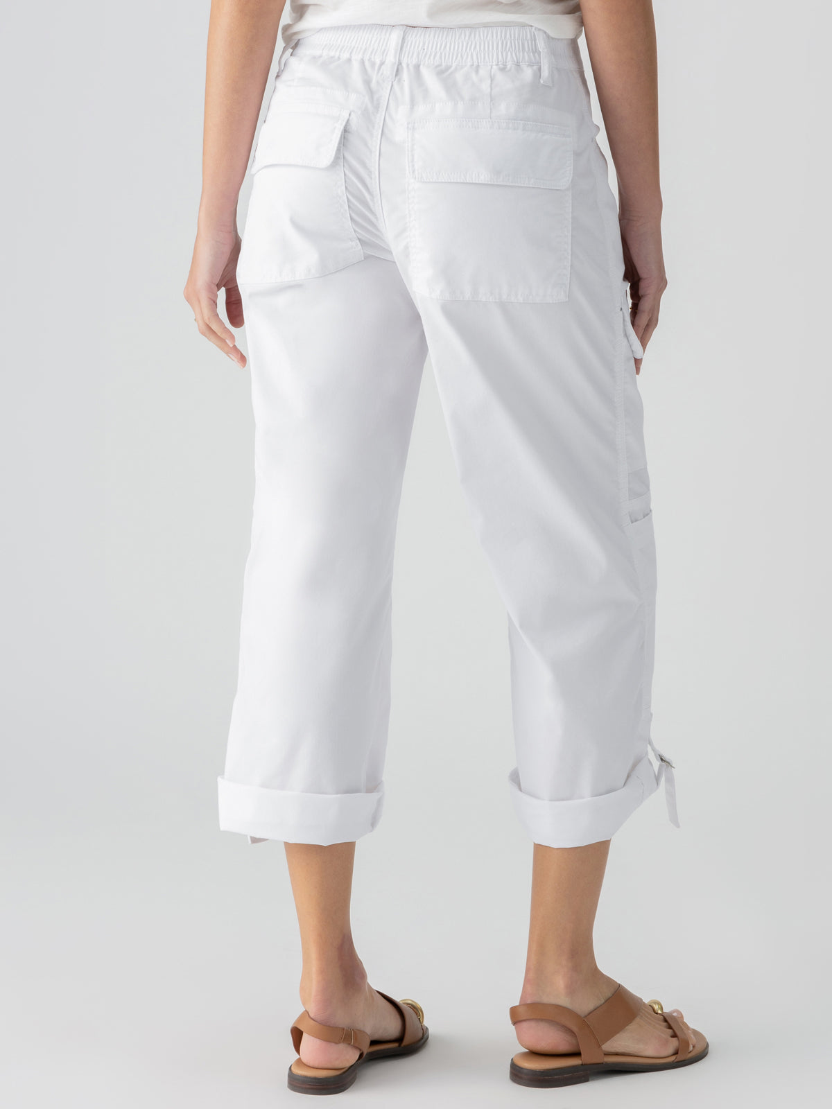 A person is shown from the waist down, wearing the white Cali Cargo Pant by Sanctuary Clothing. The slightly loose-fitting, cropped pants feature rolled cuffs and large back pockets. They are also wearing brown sandals against a plain, light-colored background.