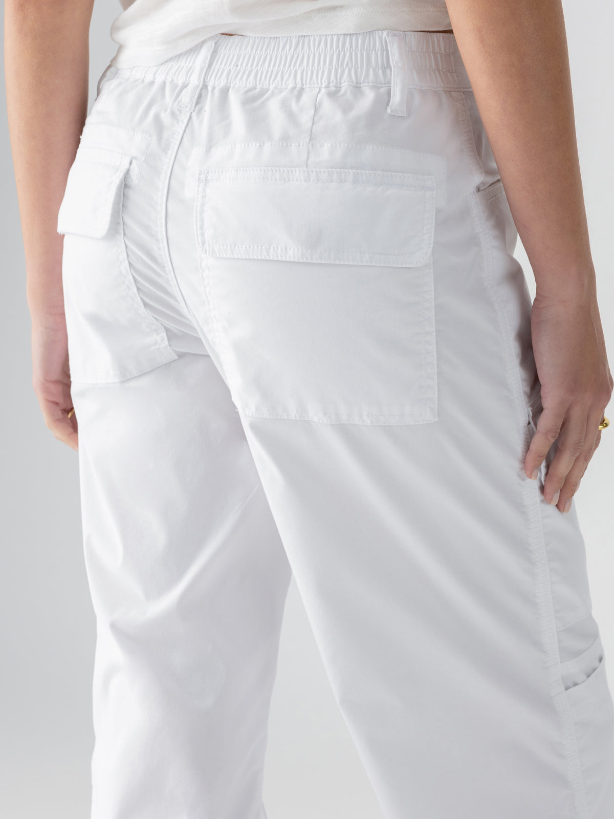 Close-up of a person wearing the Cali Cargo Pant White by Sanctuary Clothing. The pants feature an elastic waistband and two large back pockets with flaps. The person is standing with their back to the camera, casually resting one hand on their side, revealing a glimpse of a white shirt above.