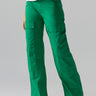 A person is wearing the Sanctuary Clothing 'Low Slung Y2K Pant Jelly Bean' in a bright green shade, paired with a white top, partially visible from the waist down. The pants feature multiple pockets and the person poses with one hand on their hip. They are also wearing open-toe sandals.