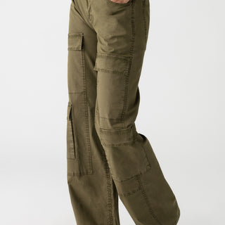 A person is shown from the waist down, wearing the Low Slung Y2K Mossy Green cargo pants from Sanctuary Clothing, featuring multiple pockets and black ballet flats. The pants have a loose fit with a button closure at the waistband against a plain white background.