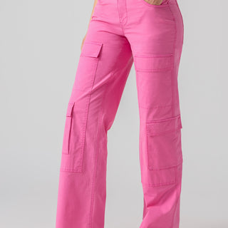 A person is wearing the Low Slung Y2K Wild Pink cargo pants by Sanctuary Clothing, paired with a white top. They are standing with one hand in their pocket and their feet slightly apart, sporting black sandals with a gold buckle against a plain gray background.