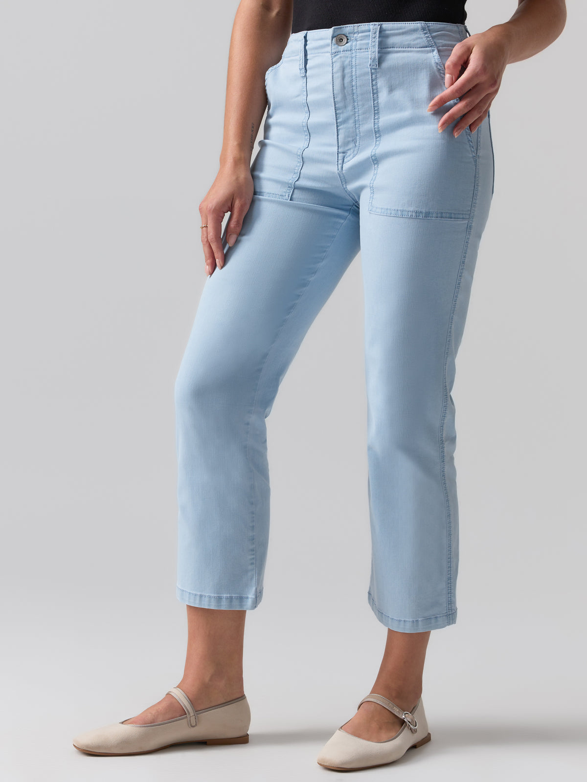 A person stands against a plain background wearing Sanctuary Clothing's Vacation Crop Pant Ultra Pale, a pair of light blue, high-waisted, straight-leg jeans with a front seam detail and a black top. They are also wearing beige flats with ankle straps. The person's hands rest casually on their thighs.