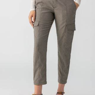 A person is wearing the Polished Cargo Compact Houndstooth pants from Sanctuary Clothing, which are high-waisted with pocket flaps, paired with a tucked-in white lace top. They complete the look with black, pointed-toe flats featuring ankle straps. The background is plain and light-colored.