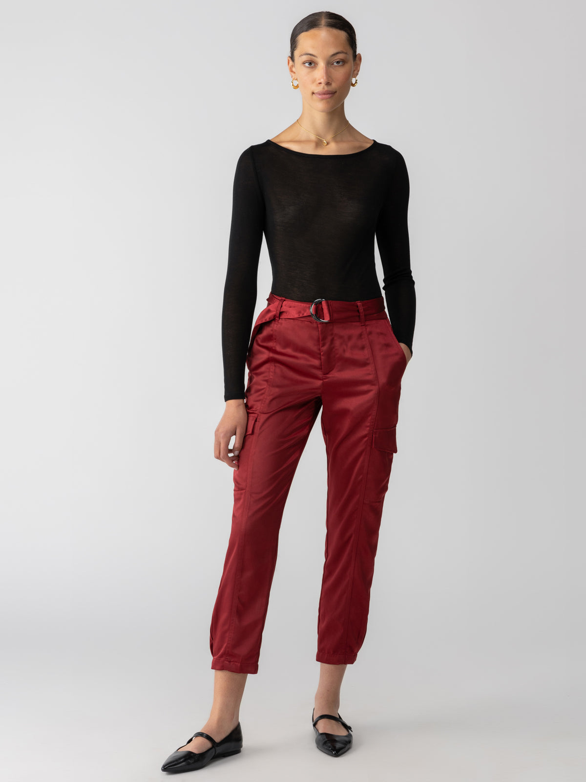 A person with long, tied-back hair is wearing a long-sleeved black top and Sanctuary Clothing's Classy Cargo Trouser Garnet with a belt, paired with black flats. They are posing against a plain white background, one hand in their pocket and the other relaxed by their side.