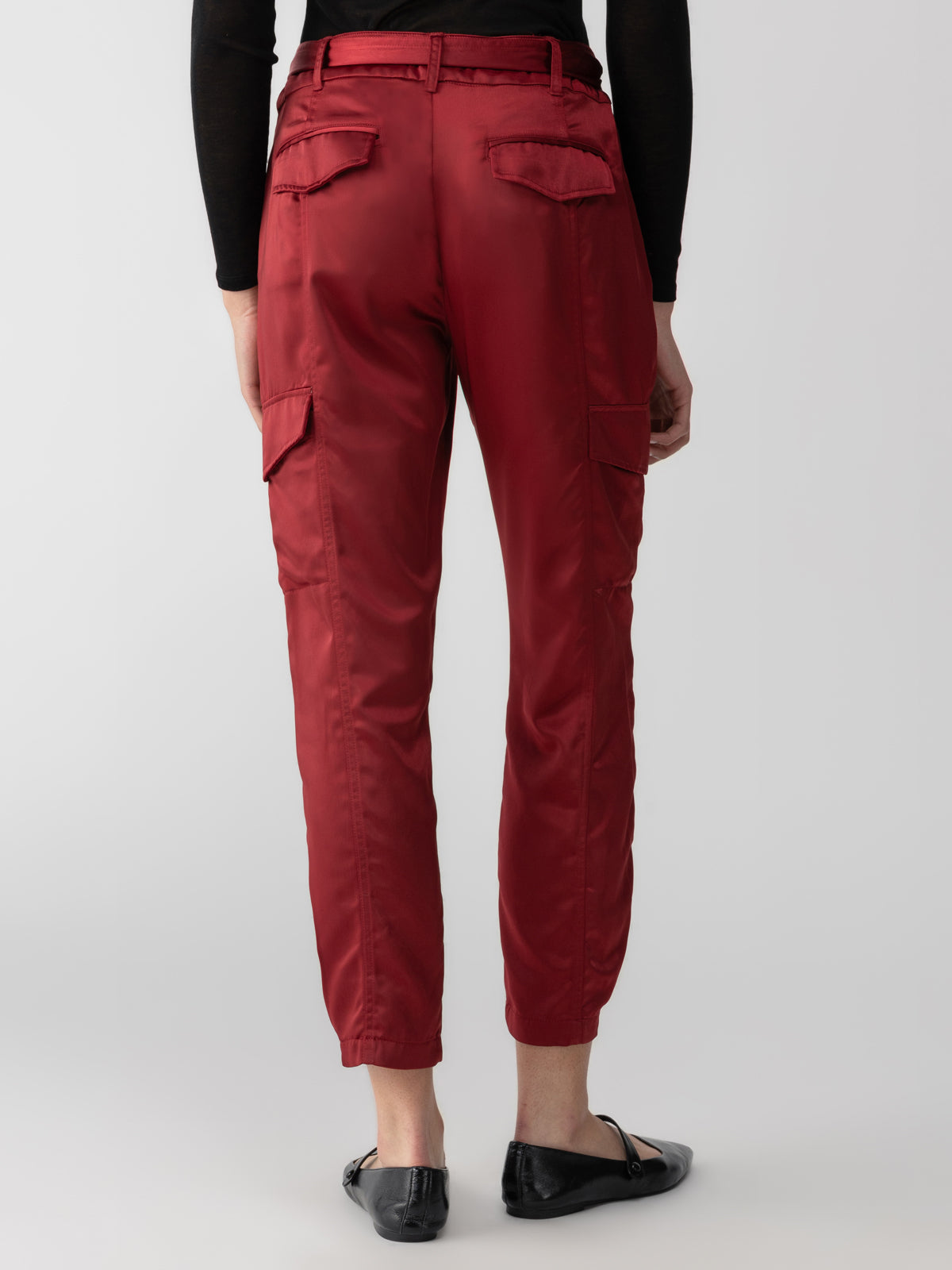 A person is standing with their back to the camera, wearing Sanctuary Clothing's Classy Cargo Trouser Garnet and black shoes. The trousers feature multiple pockets on the back and sides. The person is also wearing a black long-sleeve top against a plain, light gray background.