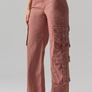 A person wearing the Y2K Strappy Pant Ash Rose by Sanctuary Clothing, featuring multiple pockets and metal rings on the sides. The pants are paired with light-colored footwear. The person is standing against a plain, light gray background, with only the lower half of their body visible.