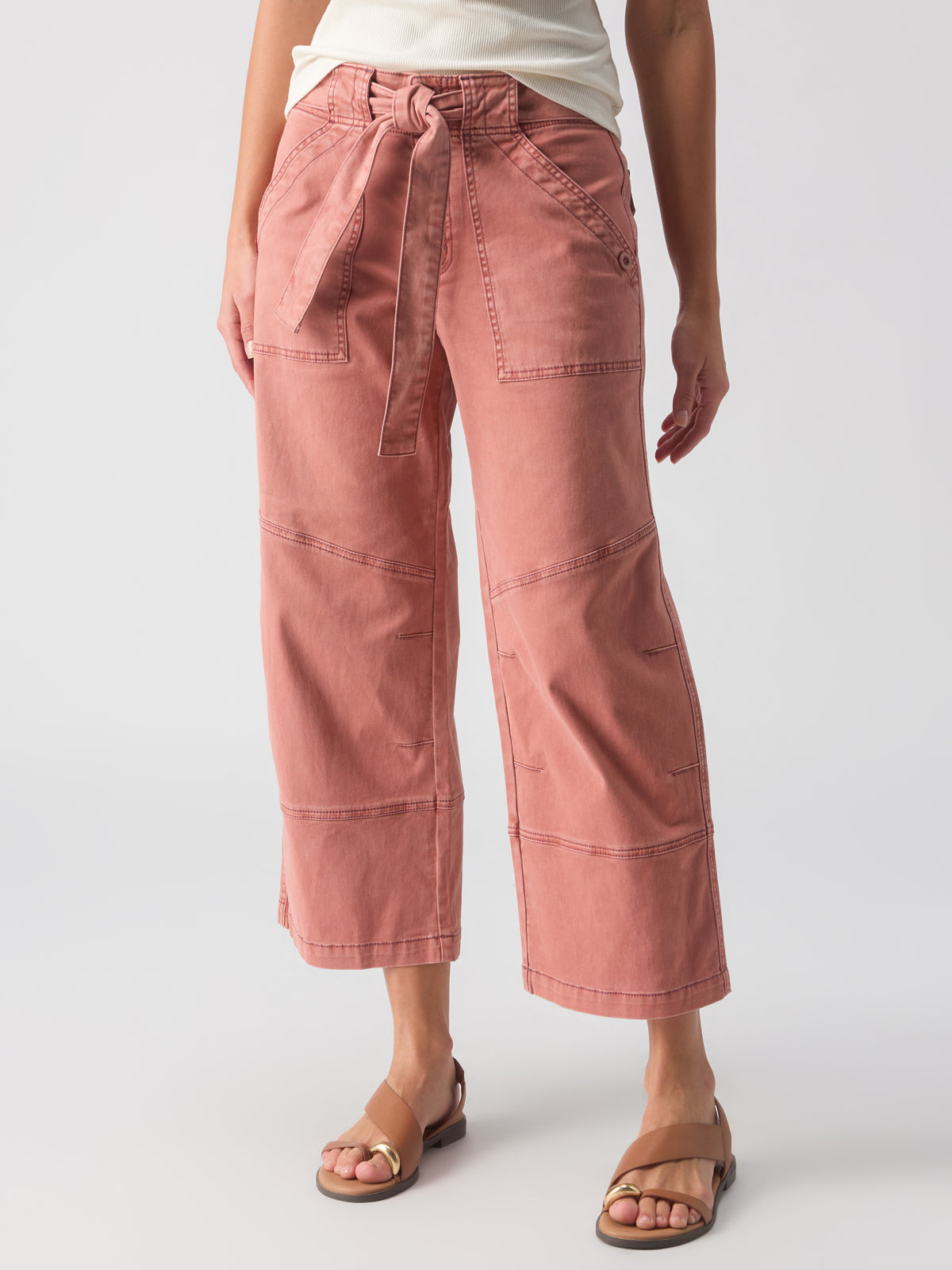 A person is seen from the waist down wearing Sanctuary Clothing's Reissue 90S Sash Ash Rose, high-waisted, wide-leg pink trousers with large front pockets and a tied belt. They are also wearing a white top and brown sandals against a plain white background.