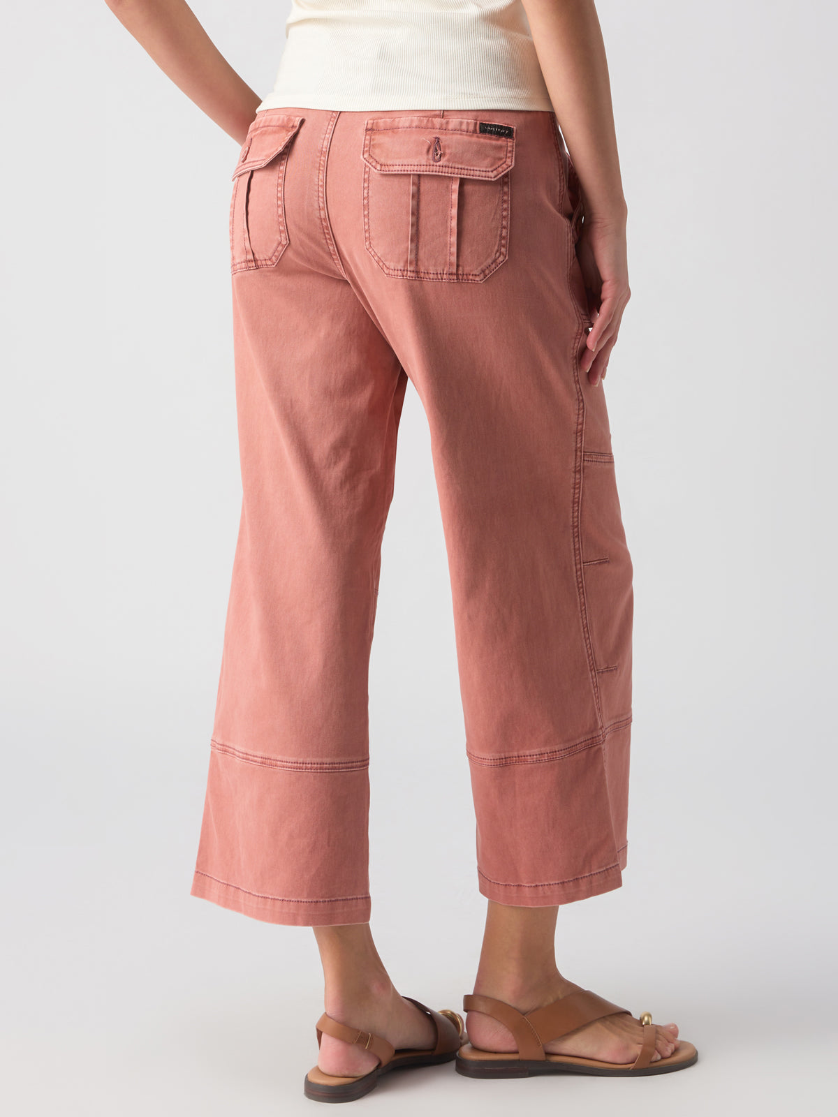 A person is standing with their back turned, wearing a white top paired with Sanctuary Clothing's Reissue 90S Sash Ash Rose pants. The loose, cropped peach-colored pants feature multiple pockets, visible stitching, and a relaxed fit. They complete the look with tan sandals against a plain, light-colored background.