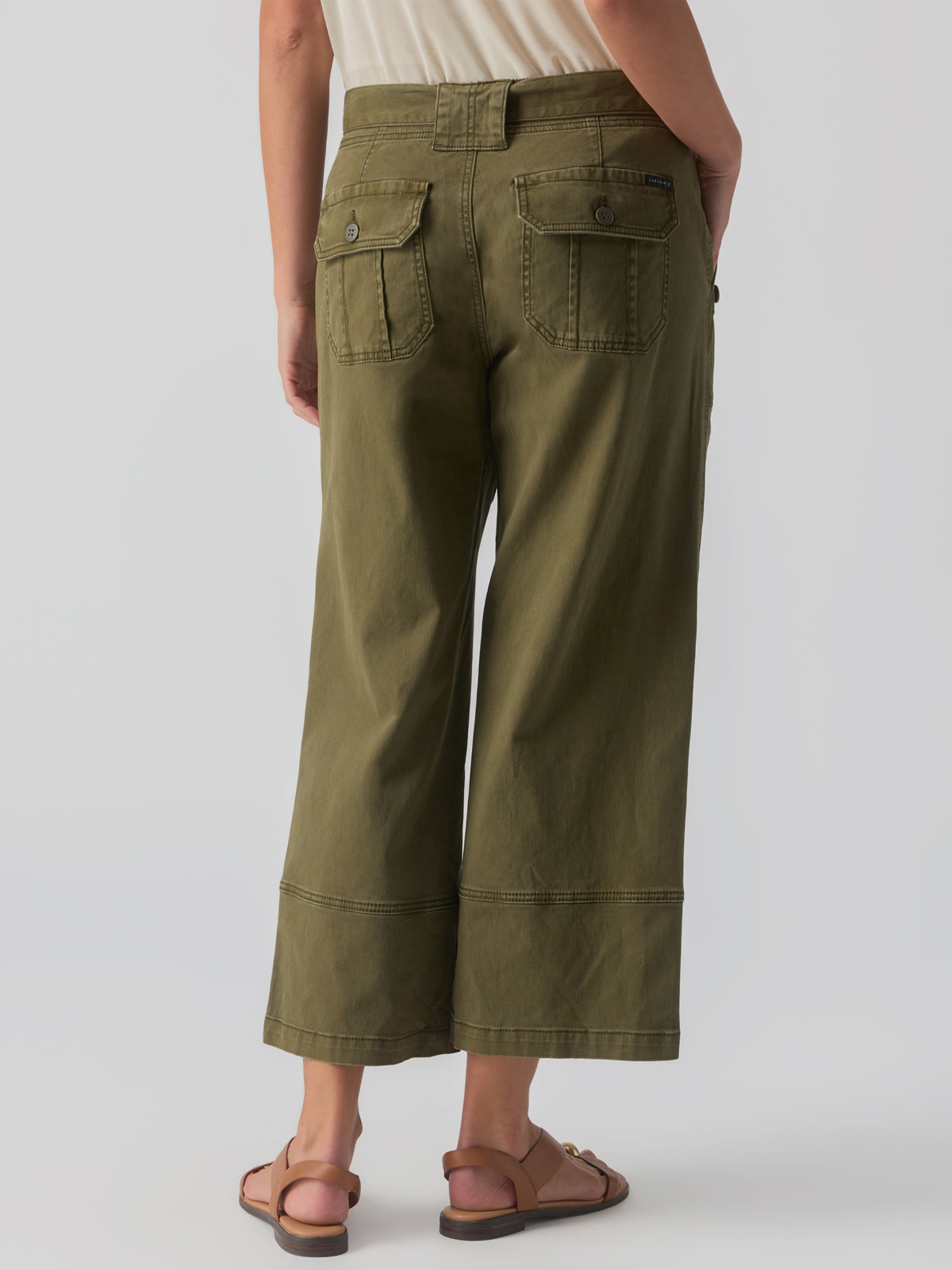 A person stands facing away from the camera, wearing Sanctuary Clothing's Reissue 90S Sash Burnt Olive wide-leg cargo pants with button-flap pockets and a beige top. They are also wearing tan sandals. The background is plain and light-colored.