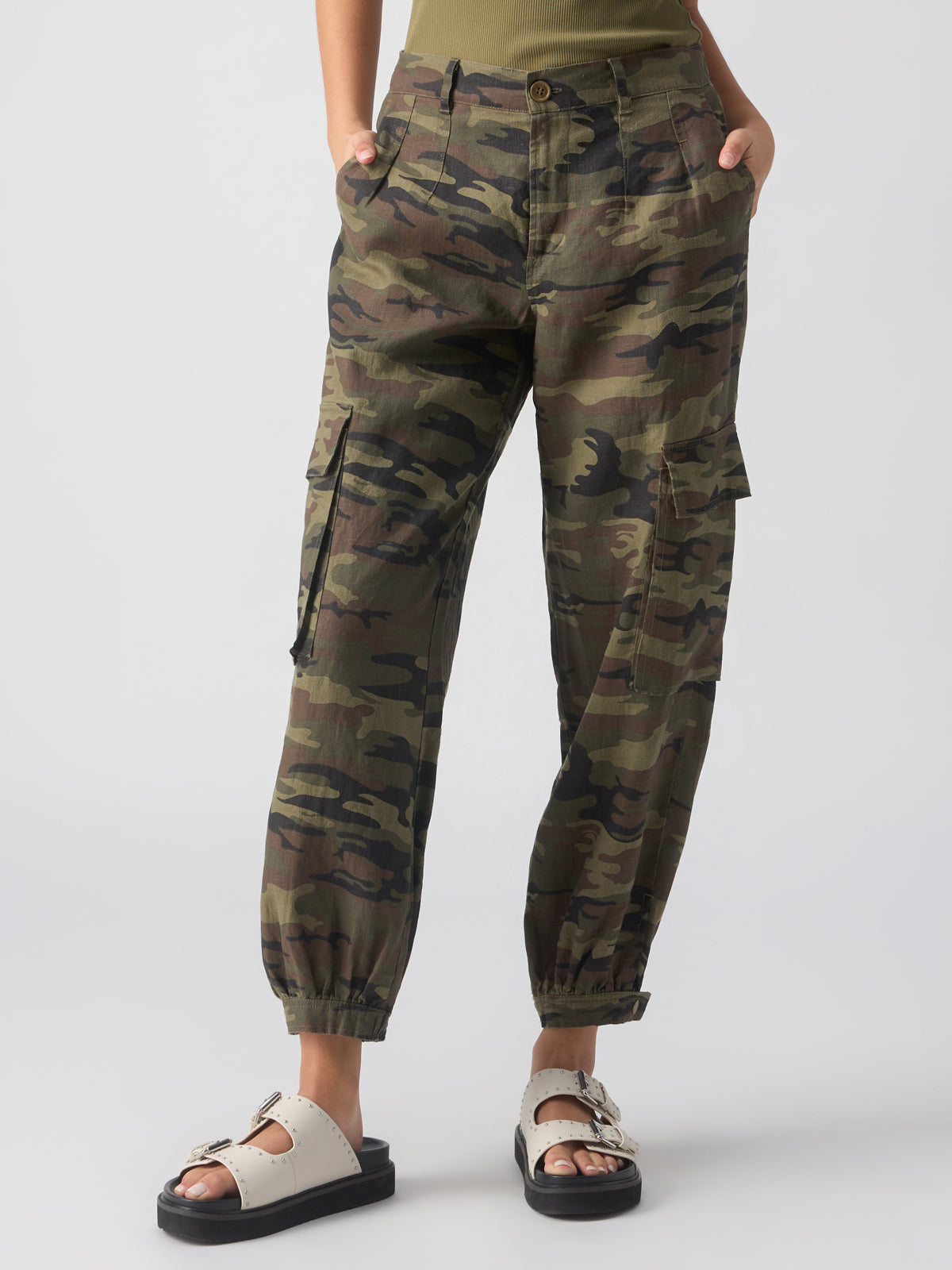 A person is standing with hands in their pockets, wearing the Pleated Linen Cargo Little Hero Camo pants from Sanctuary Clothing and white sandals featuring two buckles on each foot. They are dressed in a casual outfit against a plain white background.