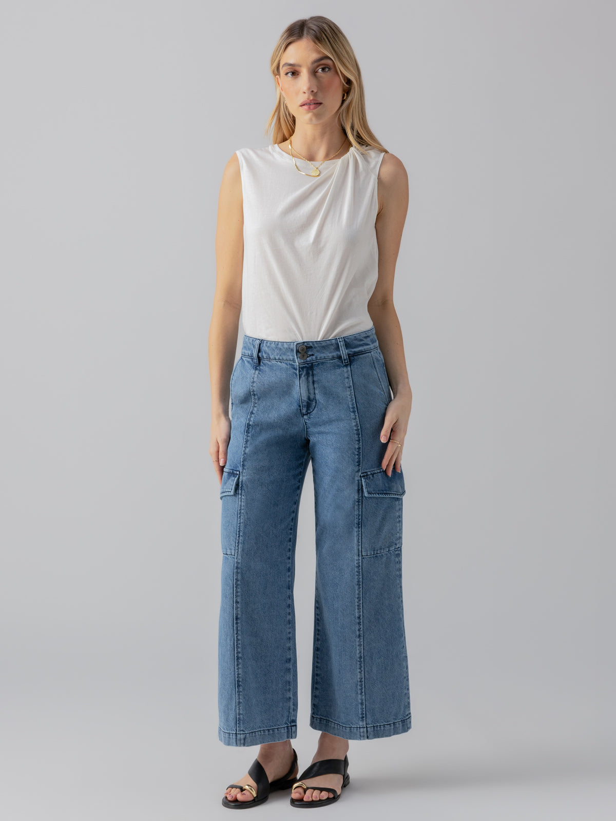 A woman with long blonde hair is standing against a plain background. She is wearing a sleeveless white top, the Rebel Wide Leg Crop Pant in Hazy Blue from Sanctuary Clothing, and black open-toe sandals. She has a neutral expression and her arms are relaxed at her sides.