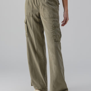 A person wearing the Relaxed Reissue Burnt Olive cargo pants by Sanctuary Clothing and a colorful shirt stands with one hand by their side. Their feet are clad in white sandals with black soles. The background is plain gray.