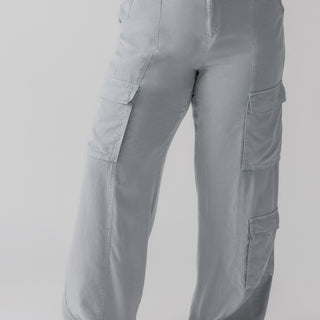 A person is wearing the Doheny Cargo Eucalyptus pants from the Inclusive Collection by Sanctuary Clothing and a white top. They are standing in a neutral pose, showing the front of the pants, and are wearing black flat shoes. The background is plain and gray.