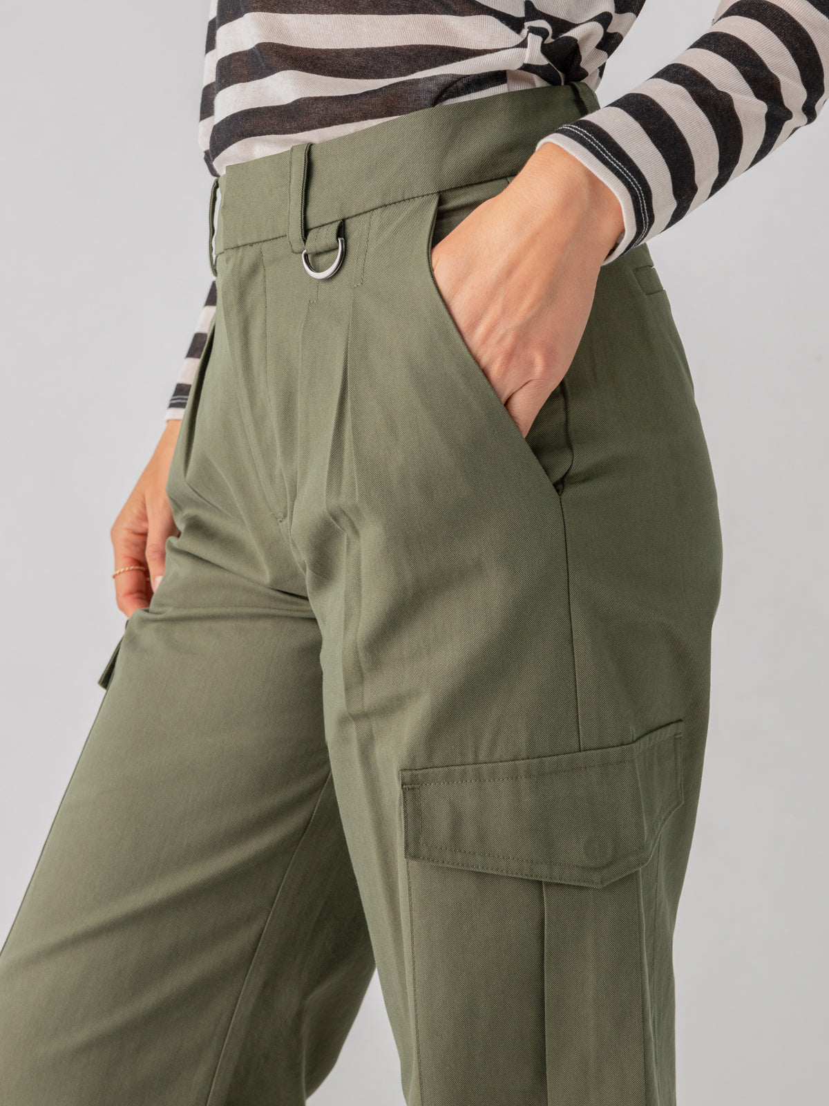 A person wearing a black and white striped long-sleeve shirt paired with the Frankie Cargo Dark Olive pants by Sanctuary Clothing, with hands in the pockets. The pants feature a belt loop with a metal ring and large side pockets. The background is plain and neutral.