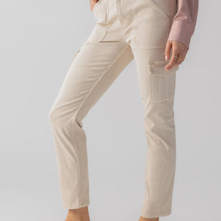 A person stands angled sideways, showcasing the Sculpted Hayden Cargo Toasted Almond pants by Sanctuary Clothing. The beige high-waisted straight-leg pants feature large front pockets and side cargo pockets. They wear a dusty pink long-sleeve blouse tucked into the pants and beige flat shoes with bows. The background is a solid light color.