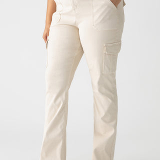 A person wearing the Sculpted Hayden Bootcut Pants in Toasted Almond from the Sanctuary Clothing Inclusive Collection and a black top has one hand in their pocket. They are also wearing black pointed-toe shoes, standing against a plain white background.