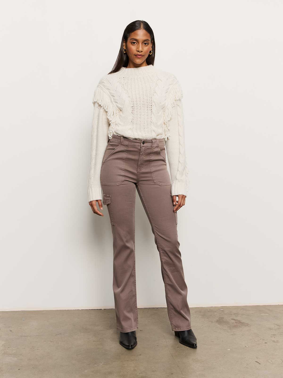 A person stands against a plain white wall, wearing the Crafted Fringe Sweater Chalk by Sanctuary Clothing and taupe high-waisted pants. They have long dark hair and are wearing black shoes, with a confident expression.