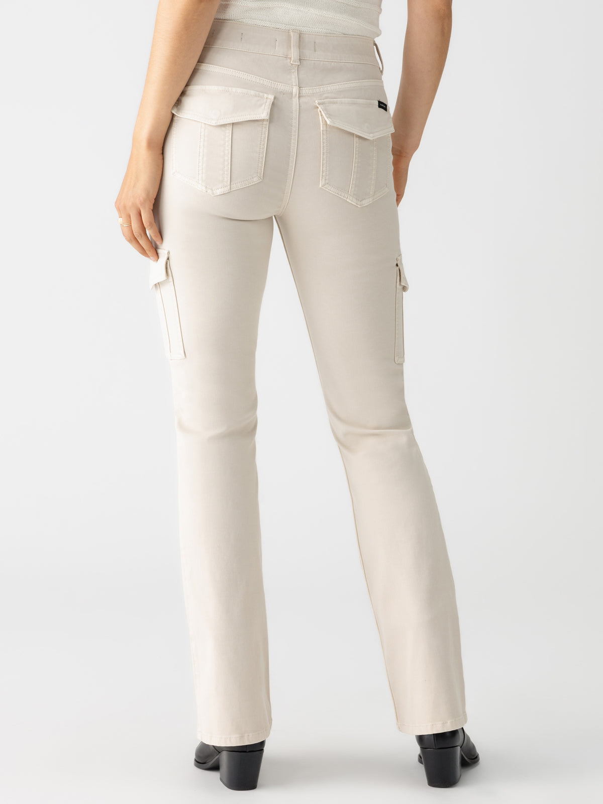 A person standing with their back to the camera, wearing Sanctuary Clothing's Sculpted Hayden Bootcut jeans in Toasted Almond. The jeans feature multiple pockets and light stitching details, with a bootcut leg that complements the black heeled shoes. The person is also wearing a white top.