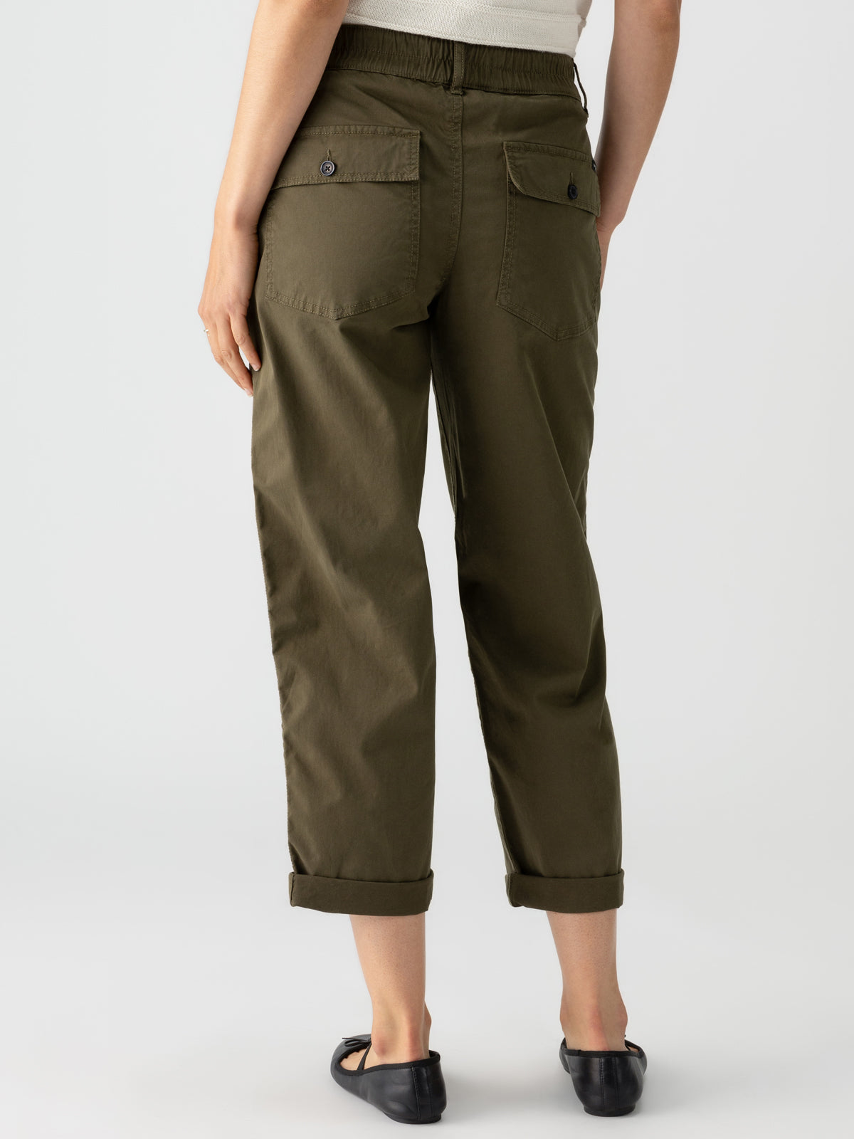 A person is shown from the back, wearing Sanctuary Clothing's Cruiser Chino in Burnt Olive, featuring ankle-length pants with rolled-up cuffs and two buttoned pockets. The person is also wearing black flat shoes and a light-colored top. The background is plain and light-colored.