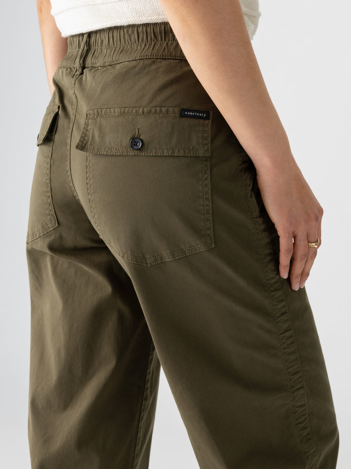 A close-up of the back of a person wearing Sanctuary Clothing's Cruiser Chino Burnt Olive pants. The pants feature a buttoned back pocket and an elastic waistband. The person's hand, adorned with a gold ring, rests by their side against a plain, light background.