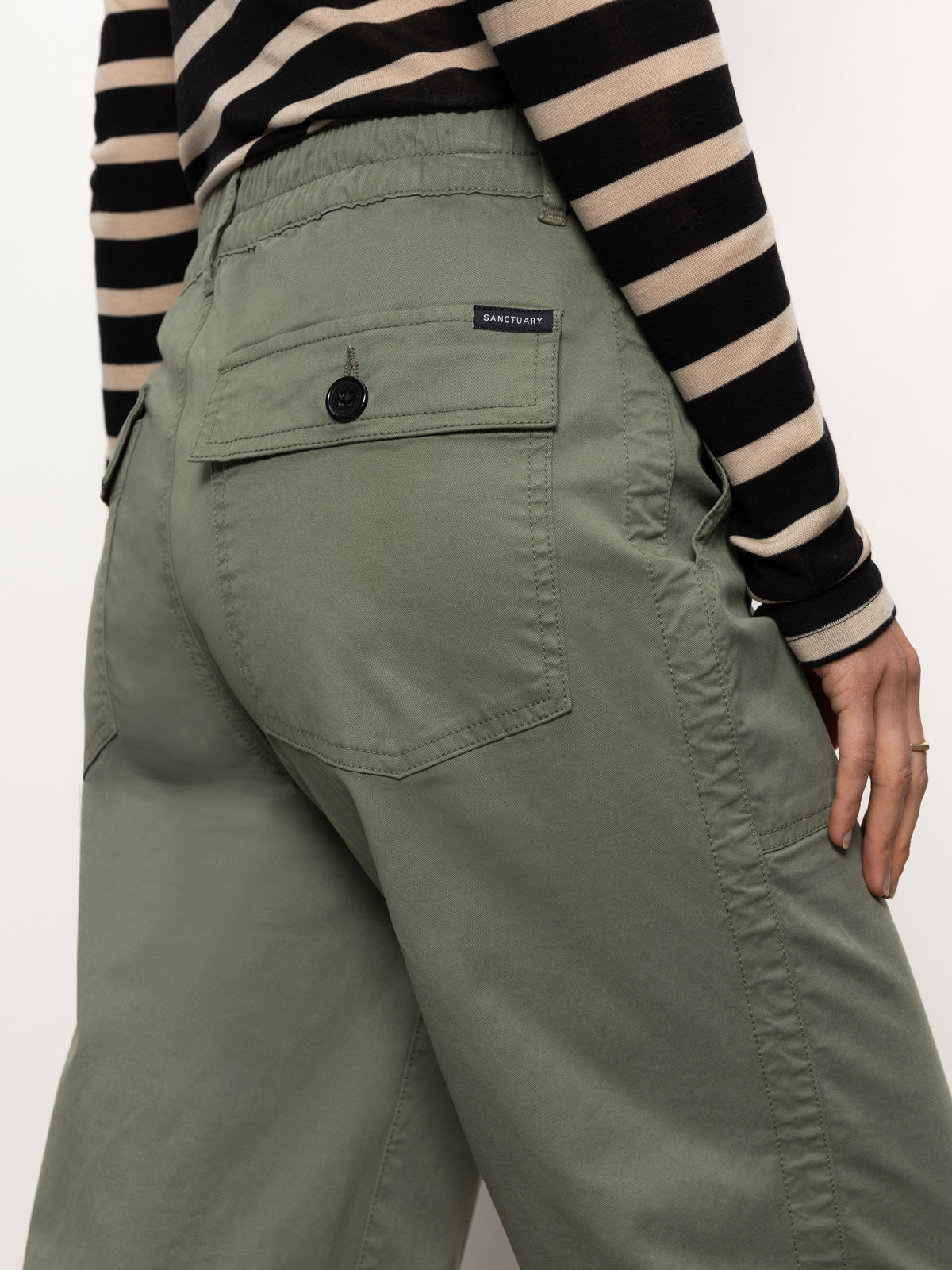 A person wearing Cruiser Chino Dark Spruce by Sanctuary Clothing pairs them with a black and beige striped long-sleeve shirt. The focus is on the back pocket with a brand label, and a hand rests on the hip. These pants feature large pockets and are made from soft, casual fabric.